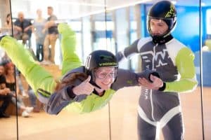 En la emocionante Experiencia en Túnel de Viento en Madrid, una persona con un traje verde y gris y casco disfruta del paracaidismo en interiores mientras un instructor le proporciona asistencia cercana. - ForTwo - Planes para parejas