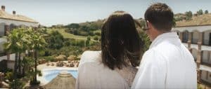 Una pareja en bata contempla la serenidad de La Cala Golf Málaga, con una impresionante piscina, exuberantes palmeras y colinas lejanas bajo un cielo despejado. Disfrute del Circuito de Hidroterapia en el Spa de La Cala Golf Málaga para una relajación total en este tranquilo paraíso. - ForTwo - Planes para parejas