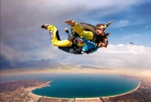 Dos personas disfrutan de un salto tándem, volando por cielos azules sobre una impresionante playa circular, experimentando un emocionante paracaidismo con las impresionantes vistas costeras de Skydive Empuriabrava. - ForTwo - Planes para parejas