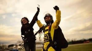 Dos paracaidistas celebran con alegría en el suelo con paracaídas y material, bajo un cielo nublado, después de un emocionante "Salto tándem de paracaidismo con Skydive Empuriabrava". - ForTwo - Planes para parejas