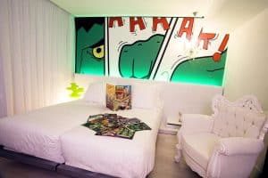 Explora la habitación con temática de cómics del Hotel Dormirdcine Madrid, que cuenta con un vibrante mural de arte pop, cómics en la cama y un elegante sillón blanco adornado. - ForTwo - Planes para parejas