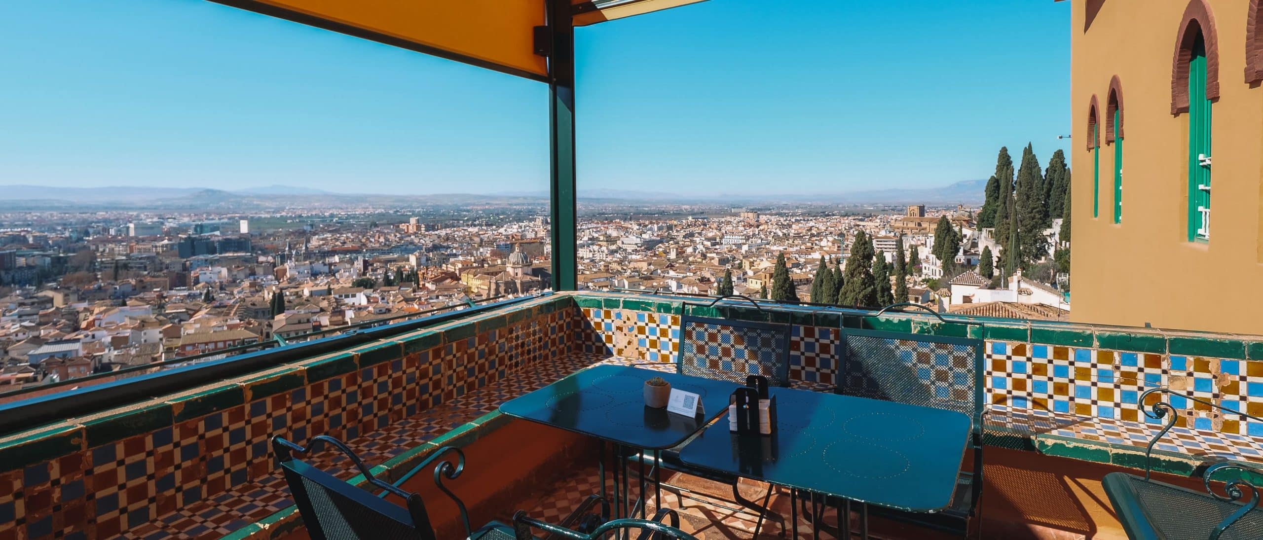 La terraza de la azotea del Hotel Alhambra Palace Granada, con azulejos de colores, ofrece una vista panorámica de la ciudad bajo un cielo azul claro, evocando el encanto del Palacio de la Alhambra. - ForTwo - Planes para parejas