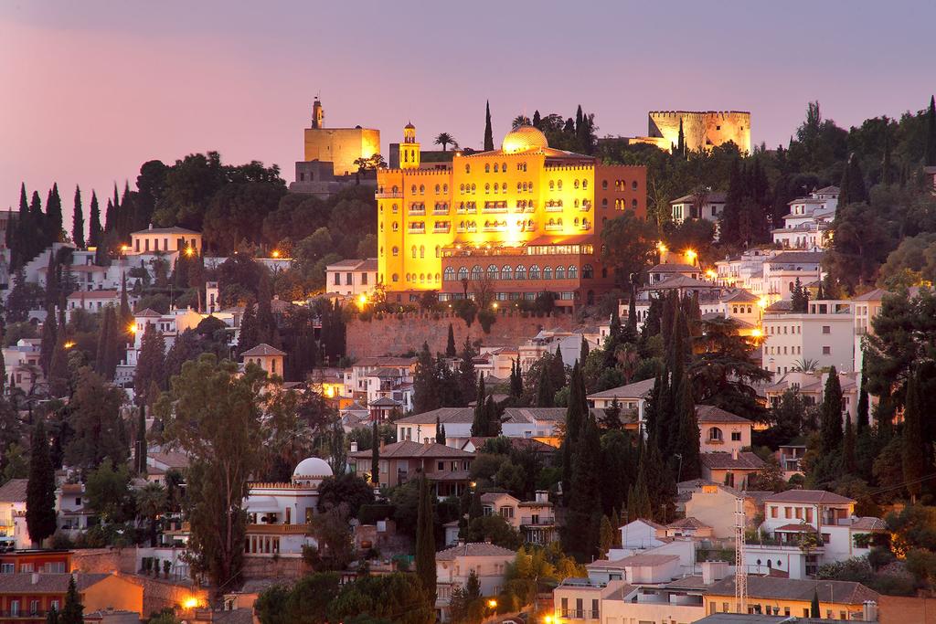 El iluminado Hotel Alhambra Palace Granada corona una ciudad en la cima de una colina al anochecer, brillando como una joya en medio de una exuberante vegetación, con el majestuoso palacio de Granada en el horizonte. - ForTwo - Planes para parejas