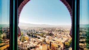 Una vista del paisaje urbano a través de una ventana arqueada en el Hotel Alhambra Palace Granada revela los tejados de Granada y las montañas distantes bajo un cielo azul claro. - ForTwo - Planes para parejas
