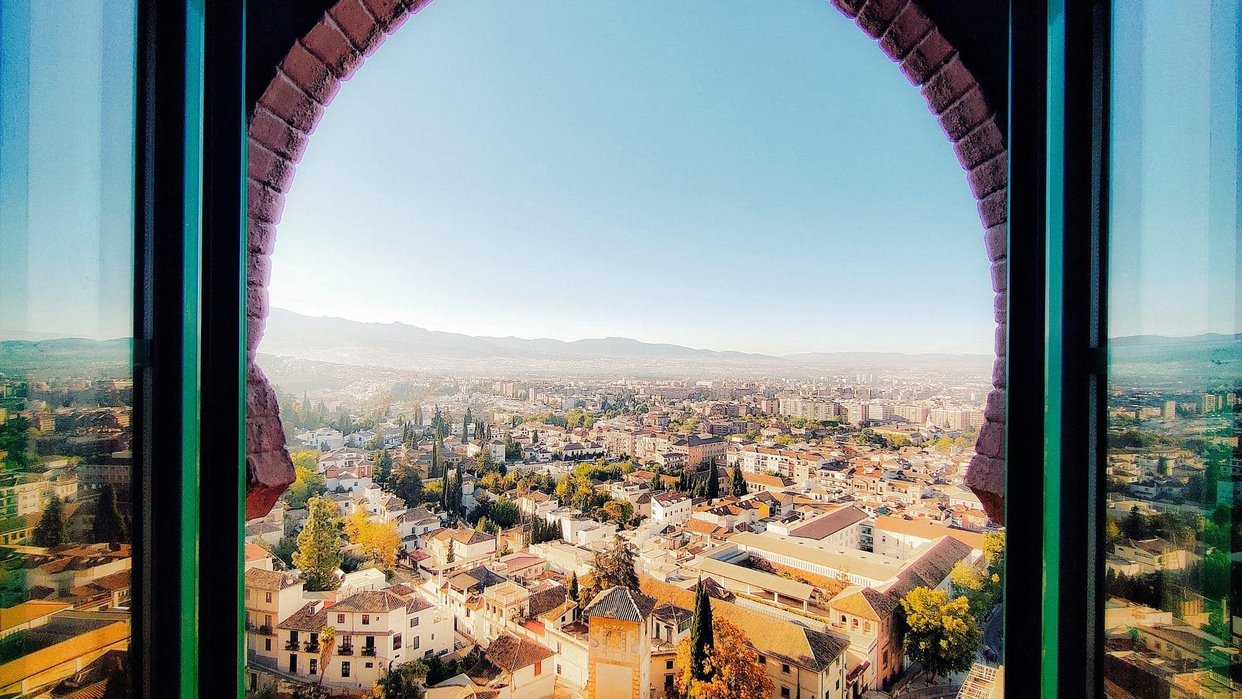 Una vista del paisaje urbano a través de una ventana arqueada en el Hotel Alhambra Palace Granada revela los tejados de Granada y las montañas distantes bajo un cielo azul claro. - ForTwo - Planes para parejas