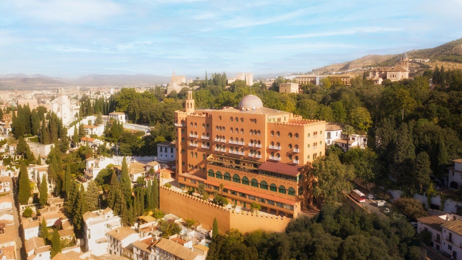 Vista aérea del Hotel Alhambra Palace Granada, una estructura de terracota rodeada de exuberante vegetación, frente a las majestuosas montañas de Granada bajo un vasto cielo azul. - ForTwo - Planes para parejas