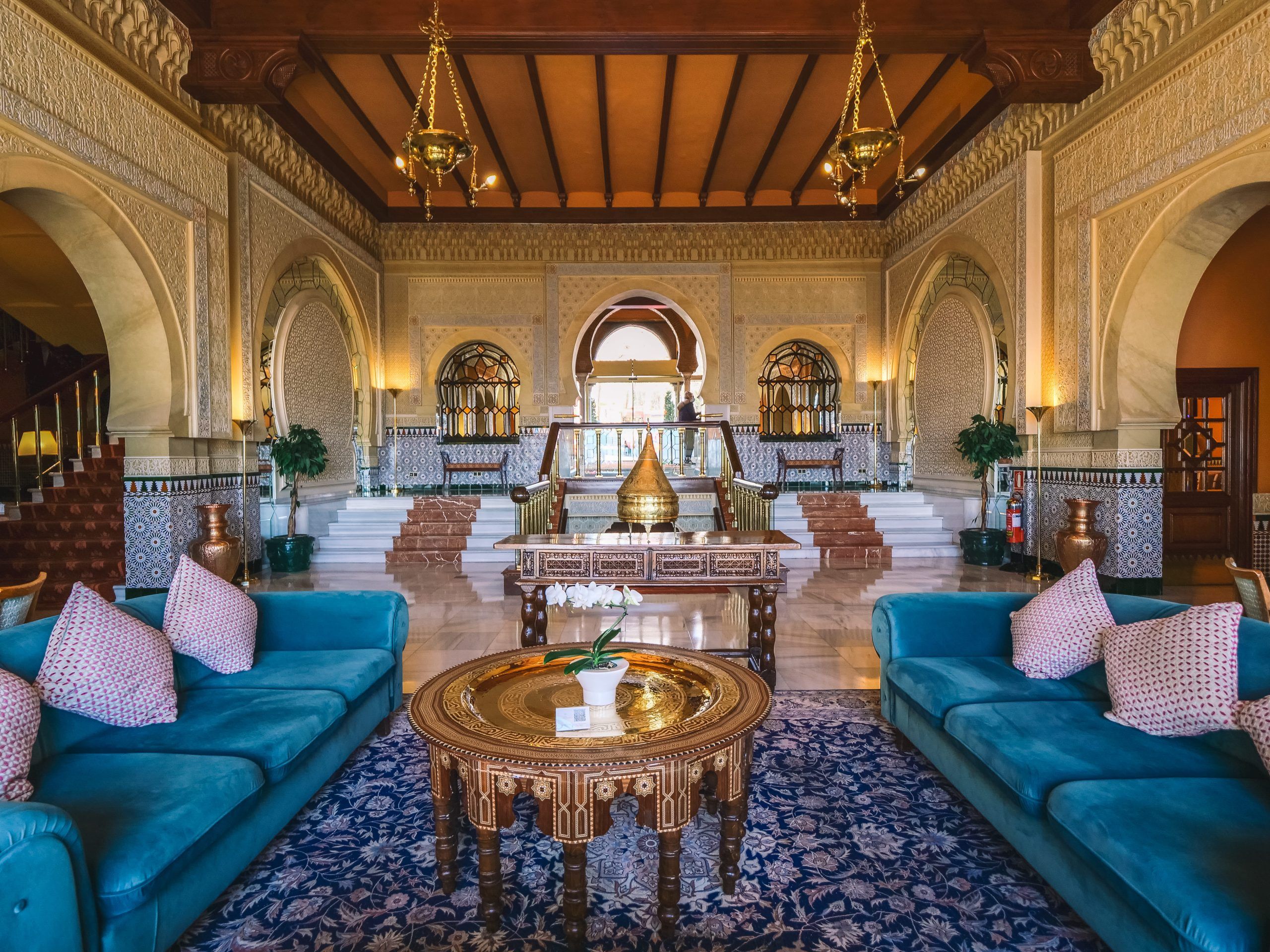 El lujoso vestíbulo del Hotel Alhambra Palace Granada cuenta con intrincados arcos inspirados en la rica historia de la ciudad, combinados con sofás azules, una mesa decorativa y elegantes candelabros. - ForTwo - Planes para parejas