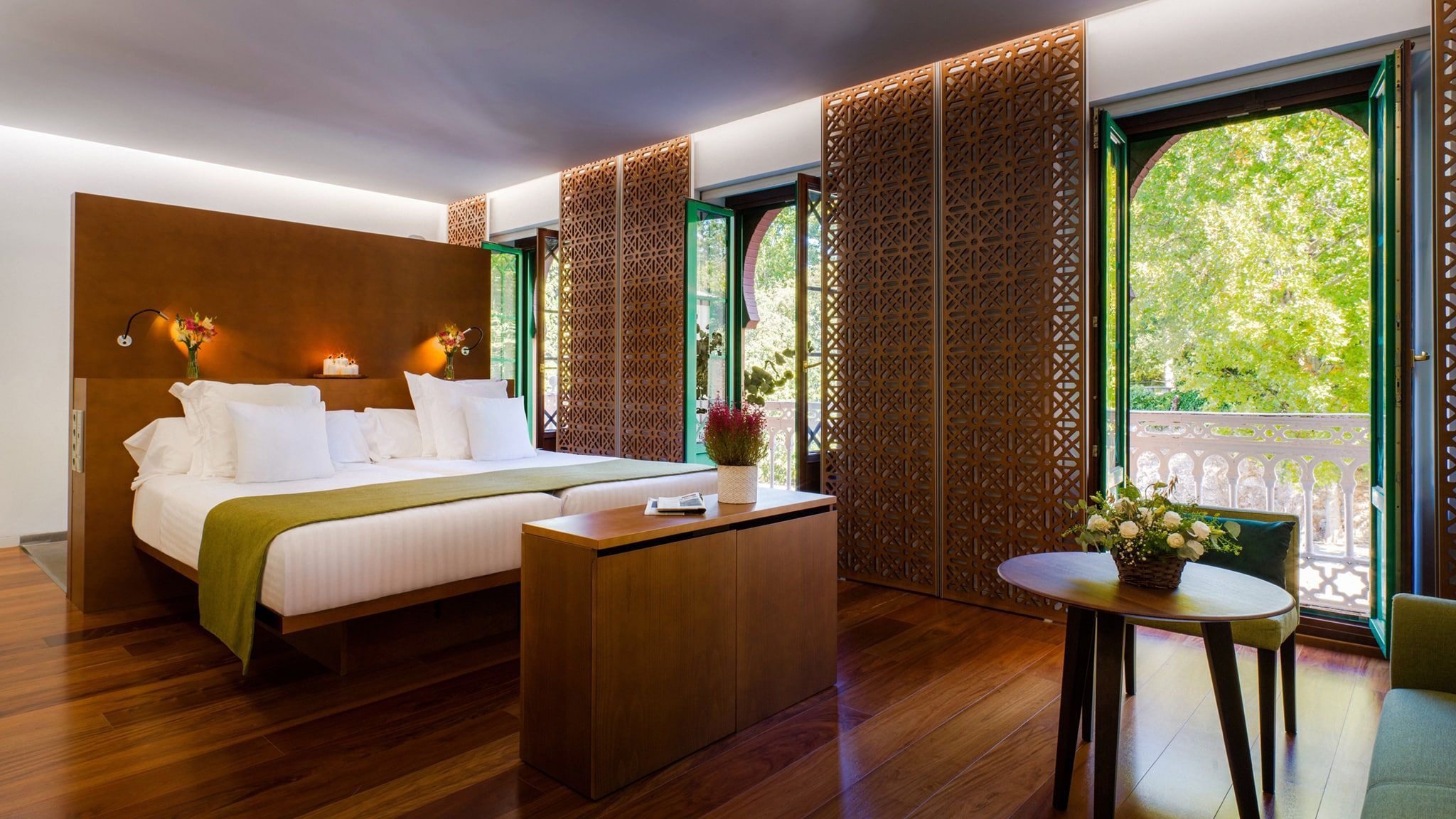 Una habitación del Hotel Alhambra Palace Granada cuenta con una cama grande, decoración de madera, plantas exuberantes y ventanas arqueadas que ofrecen impresionantes vistas a la vegetación de Granada. - ForTwo - Planes para parejas