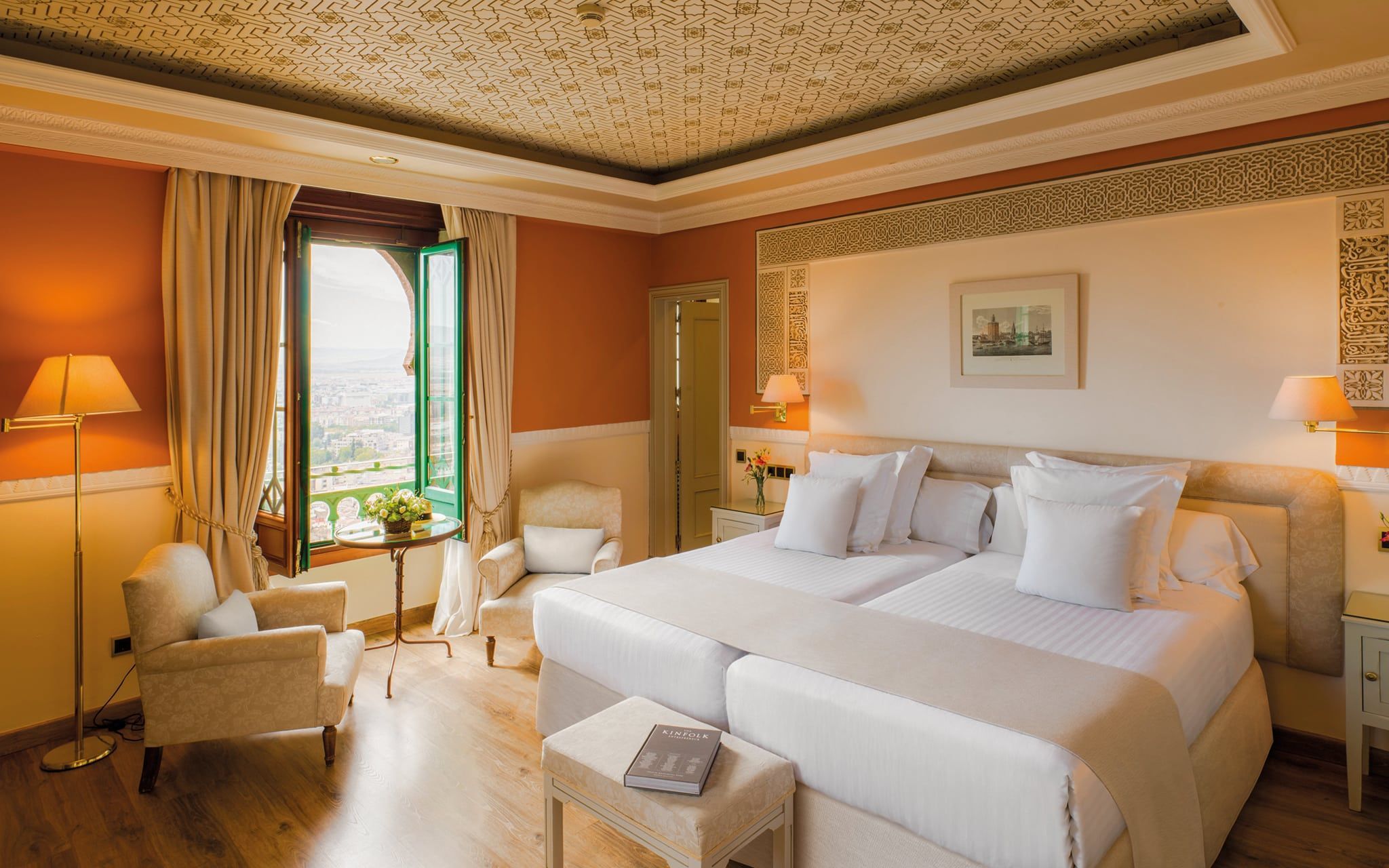 Descubra la elegancia en el Hotel Alhambra Palace Granada con una habitación bellamente diseñada que cuenta con una cama grande y dos sillas junto a la ventana, y ofrece impresionantes vistas del paisaje urbano de Granada. - ForTwo - Planes para parejas