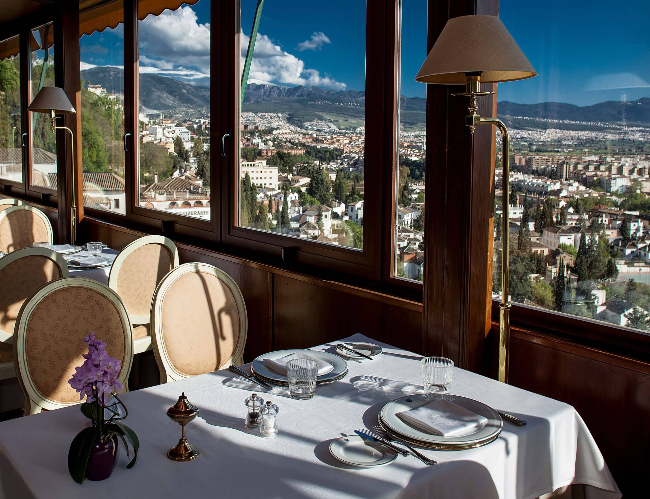 El elegante restaurante del Hotel Alhambra Palace Granada ofrece una vista panorámica de la ciudad, con mesas dispuestas con manteles blancos, cada una adornada con un centro de mesa de flores moradas y bañadas por la luz del sol. - ForTwo - Planes para parejas