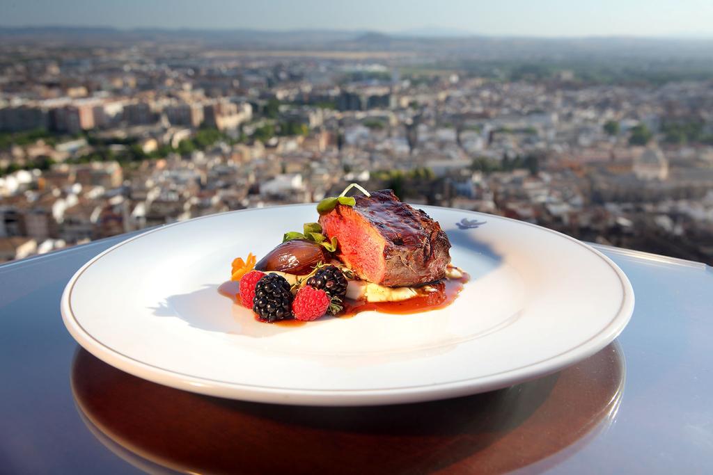 Un plato de bistec gourmet con bayas picantes adorna un plato blanco, que encarna la esencia del Hotel Alhambra Palace Granada, mientras el majestuoso paisaje urbano y el Palacio de la Alhambra brindan un impresionante telón de fondo. - ForTwo - Planes para parejas