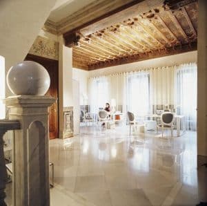 Una elegante habitación con suelos de mármol, techo de madera decorativa y muebles blancos modernos que evocan el Hotel Hospes Palacio de los Patos Granada. La luz del sol se cuela por las cortinas. - ForTwo - Planes para parejas