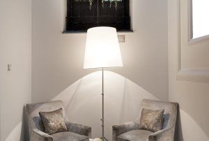 Acogedora zona de estar inspirada en el Hotel Hospes Puerta de Alcalá 5 estrellas Madrid, con dos sillones, una lámpara de pie y almohadas decorativas bajo una ventana alta adornada con exuberantes plantas, creando un lugar acogedor para relajarse como si estuviera en el centro de Madrid. - ForTwo - Planes para parejas