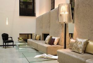 Un moderno salón en el Hotel Hospes Puerta de Alcalá 5 estrellas Madrid cuenta con altos sofás beige, mesas de cristal, almohadas decorativas, un libro abierto y una elegante lámpara de pie. - ForTwo - Planes para parejas