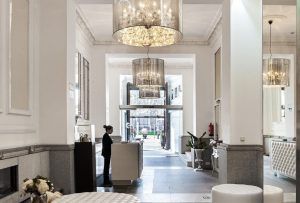 El elegante vestíbulo del Hotel Hospes Puerta de Alcalá 5 estrellas Madrid exhibe lámparas de araña, un elegante mostrador de recepción y una decoración moderna con una persona atenta detrás del mostrador. - ForTwo - Planes para parejas