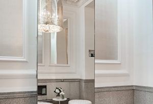 Esta elegante habitación del Hotel Hospes Puerta de Alcalá 5 estrellas de Madrid cuenta con grandes espejos, una lámpara de araña ornamentada y una pequeña otomana blanca. Molduras decorativas adornan las paredes, capturando un encanto majestuoso que evoca la cercana Puerta de Alcalá. - ForTwo - Planes para parejas