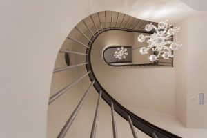La escalera de caracol del Hotel Palacete de Álamos Málaga, vista desde abajo, presenta una lámpara de araña bellamente decorativa sobre un fondo neutro. - ForTwo - Planes para parejas