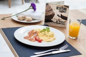 El desayuno del Hotel Palacio de Pinello Sevilla incluye huevos revueltos, beicon y tomate con zumo de naranja. Un menú y una encantadora flor realzan el acogedor ambiente. - ForTwo - Planes para parejas