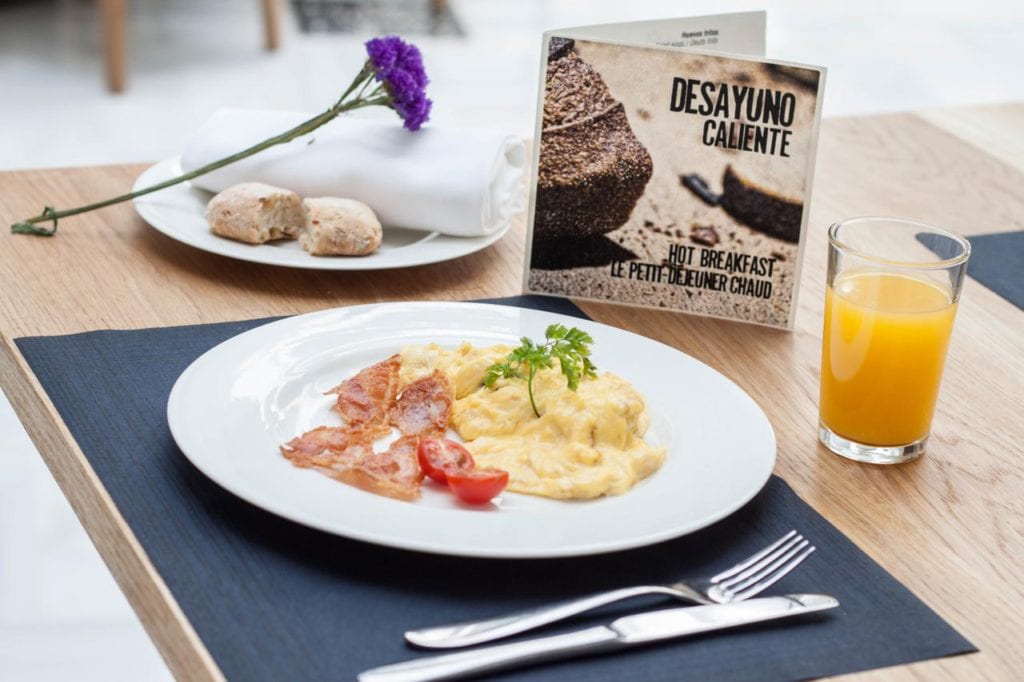 El desayuno del Hotel Palacio de Pinello Sevilla incluye huevos revueltos, beicon y tomate con zumo de naranja. Un menú y una encantadora flor realzan el acogedor ambiente. - ForTwo - Planes para parejas