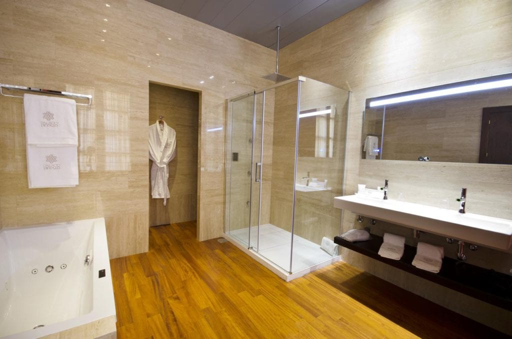 Experimente el lujo en el Hotel Palacio de Pinello Sevilla, que cuenta con un baño con suelo de madera, ducha de cristal, bañera, lavabos dobles y un elegante albornoz junto a la puerta. - ForTwo - Planes para parejas