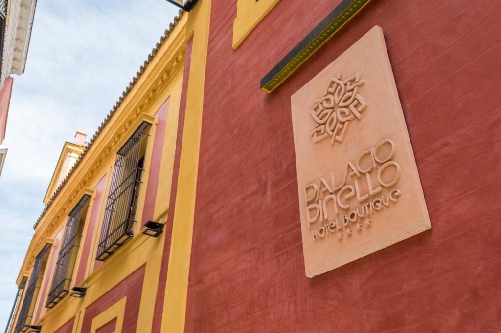 El Hotel Palacio de Pinello Sevilla presenta una llamativa fachada roja y amarilla complementada por un elegante cartel decorativo en la pared. - ForTwo - Planes para parejas