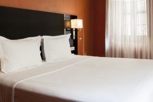 Una cama cuidadosamente hecha con sábanas blancas realza una habitación cálidamente iluminada en el Hotel Palacio de Santa Paula Granada, acentuada por paredes naranjas y cortinas transparentes, que capturan el encanto de Granada. - ForTwo - Planes para parejas