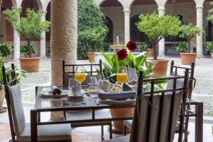 Un pintoresco patio del Hotel Palacio de Santa Paula Granada cuenta con una mesa de desayuno con flores, jugos y pasteles, rodeada de elegantes arcos y frondosos árboles en macetas. - ForTwo - Planes para parejas