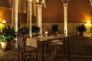 El comedor al aire libre del Hotel Palacio de Santa Paula Granada encanta con mesas y sillas en un encantador patio, rodeado de columnas iluminadas y exuberantes plantas en macetas, capturando la esencia de Granada. - ForTwo - Planes para parejas