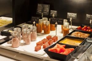 En el Hotel Palacio de Santa Paula Granada, un suntuoso buffet incluye tarros de paté, membrillo, diversos quesos y coloridos trozos de fruta en un mostrador de servicio elegantemente dispuesto. - ForTwo - Planes para parejas