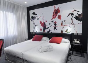 Habitación moderna en el Hotel Santo Domingo Madrid con dos camas individuales, almohadas rojas, arte abstracto en la pared, iluminación suave y una cortina blanca a la izquierda. - ForTwo - Planes para parejas