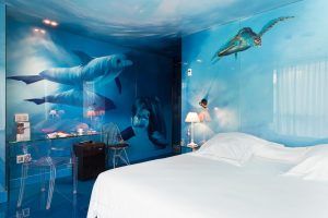 Esta habitación con temática oceánica, que recuerda a un tranquilo refugio en el Hotel Santo Domingo Madrid, cuenta con cautivadores murales de pared de delfines y tortugas marinas, complementados con una acogedora cama, un escritorio elegante y sillas transparentes. - ForTwo - Planes para parejas