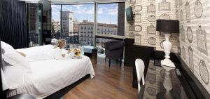 En el Hotel Santo Domingo Madrid, una acogedora habitación con vistas a la animada plaza madrileña. Una mujer se relaja en la cama con una bandeja de desayuno, complementada con la comodidad de un escritorio, una lámpara y un televisor. - ForTwo - Planes para parejas