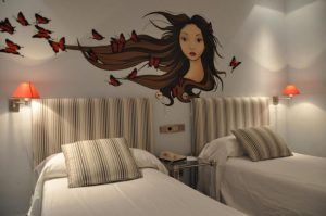 Camas individuales debajo de un impresionante mural de una mujer con cabello suelto y mariposas rojas adornan las paredes del Hotel Santo Domingo Madrid. - ForTwo - Planes para parejas