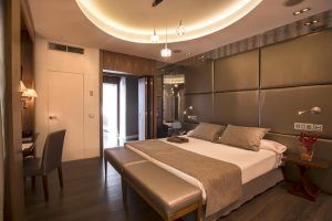 El Hotel Santo Domingo Madrid ofrece una habitación moderna en Madrid con una cama grande, iluminación tenue y un diseño elegante. Cuenta con suelos de madera oscura y detalles metálicos que le dan un toque contemporáneo. - ForTwo - Planes para parejas