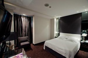 Experimente un ambiente acogedor en la moderna habitación del Hotel Santo Domingo Madrid, que cuenta con una cama doble, detalles oscuros, una silla y una TV con iluminación suave. - ForTwo - Planes para parejas