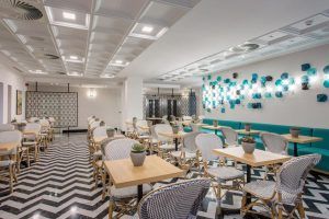 El Hotel Room Mate Valeria Málaga cuenta con una moderna cafetería con suelo de baldosas en zigzag, sillas de mimbre y mesas de madera. Una pared decorada con arte abstracto azul realza su vibrante elegancia. - ForTwo - Planes para parejas