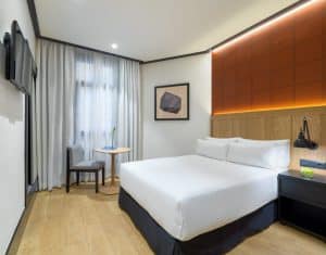 Descubra el encanto moderno de una habitación en el Hotel H10 Puerta de Alcalá Madrid, que ofrece una cama grande, un televisor de pared, un cómodo sillón y una mesa. Las obras de arte y la cálida iluminación alrededor del cabecero crean un ambiente sereno. - ForTwo - Planes para parejas