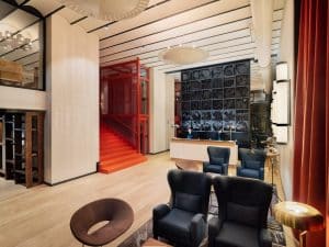 El elegante lobby del Hotel H10 Puerta de Alcalá Madrid captura su esencia con una escalera roja, sillas modernas, una galería de arte en la pared y una iluminación cálida. - ForTwo - Planes para parejas