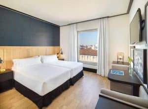 Descubra la elegancia contemporánea en nuestra habitación del Hotel H10 Puerta de Alcalá Madrid con dos camas individuales, TV de pared, un gran ventanal con impresionantes vistas a la ciudad y una decoración minimalista. - ForTwo - Planes para parejas