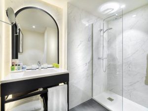 Este moderno baño, que recuerda el lujo del Hotel H10 Puerta de Alcalá Madrid, cuenta con elegantes paredes de mármol, una elegante ducha de cristal, un espejo iluminado, un lavabo con artículos de tocador y un elegante toallero. - ForTwo - Planes para parejas