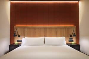 Disfrute de la comodidad y el estilo en el Hotel H10 Puerta de Alcalá Madrid con una moderna habitación que cuenta con una cama grande contra una pared de acento rojo, realzada por un cabecero de madera y dos elegantes lámparas de pared negras. - ForTwo - Planes para parejas