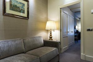 Una acogedora sala de estar cuenta con un sofá gris, una lámpara y un cuadro, que irradia el elegante encanto del Hotel Wellington Madrid. Disfrute de la vista a otra habitación a través de una puerta abierta que refleja el sofisticado estilo madrileño. - ForTwo - Planes para parejas