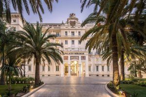 La entrada del Gran Hotel Miramar Málaga cuenta con palmeras, una puerta arqueada y el cartel del Palacio Miramar iluminado al anochecer. - ForTwo - Planes para parejas