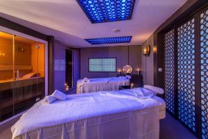 Una serena sala de spa en el Gran Hotel Miramar Málaga incluye dos mesas de masaje, iluminación tenue y un diseño moderno con relajantes detalles en azul y naranja. - ForTwo - Planes para parejas