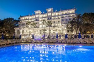 Al anochecer, el Gran Hotel Miramar Málaga brilla con una gran fachada, destacando en primer plano una radiante piscina, tumbonas y sombrillas azules elegantemente colocadas. - ForTwo - Planes para parejas