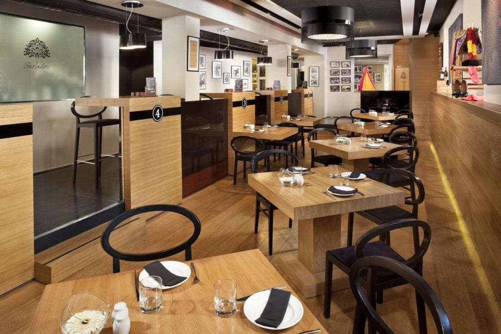 Este moderno restaurante del Hotel Gran Meliá Colón Sevilla cuenta con mesas de madera, sillas negras y una decoración minimalista. Las paredes están adornadas con fotos enmarcadas, combinando sofisticación y comodidad en un ambiente hotelero con mucho encanto. - ForTwo - Planes para parejas