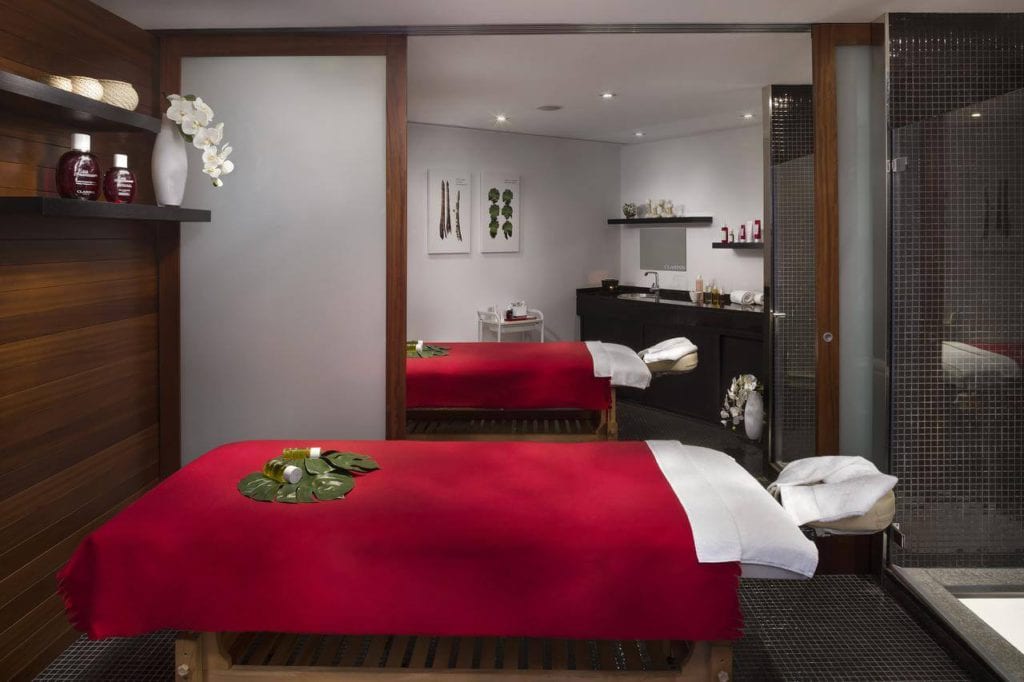 Relájese en la relajante sala de masajes del Hotel Gran Meliá Colón Sevilla con su mesa cubierta de rojo, suaves toallas blancas, elegantes orquídeas y una relajante iluminación tenue. - ForTwo - Planes para parejas