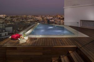 En el Hotel Gran Meliá Colón Sevilla, la piscina triangular de la azotea, al atardecer, ofrece una impresionante vista de la ciudad, perfecta para relajarse. Toallas de lujo y una cesta de frutas en la terraza de madera realzan su experiencia. - ForTwo - Planes para parejas