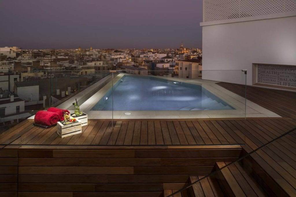 En el Hotel Gran Meliá Colón Sevilla, la piscina triangular de la azotea, al atardecer, ofrece una impresionante vista de la ciudad, perfecta para relajarse. Toallas de lujo y una cesta de frutas en la terraza de madera realzan su experiencia. - ForTwo - Planes para parejas