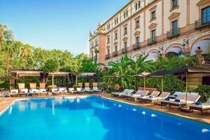 La piscina al aire libre, rodeada de tumbonas y sombrillas, ofrece una escapada tranquila con el majestuoso Hotel Alfonso XIII Sevilla como telón de fondo, enclavado entre la exuberante vegetación de Sevilla. - ForTwo - Planes para parejas